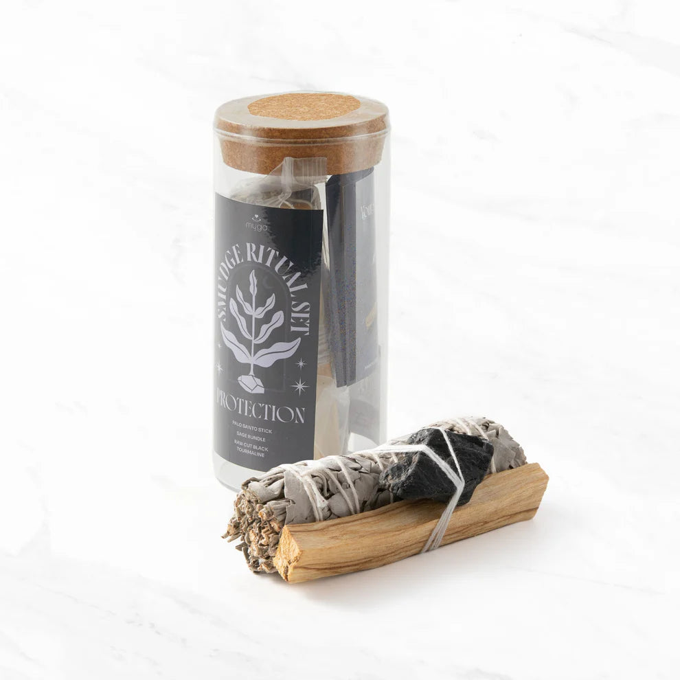 Myga Puhdistusrituaalisarja; lasipurkki tekstillä Sage Ritual Set Protection ja korkkikansi seisoo pystyssä; etiketissä yksinkertaista mustaa groteskitekstiä valkoisella paperilla; purkin vieressä nippu harmaanvihreää kuivattua salviaa ja sileä vaalea puinen palo santo -tikku sidottuna yhteen narulla; kaikki esineet lepäävät tasaisella mattavalkoisella pinnalla hajavalossa; ei ihmisiä kuvassa.