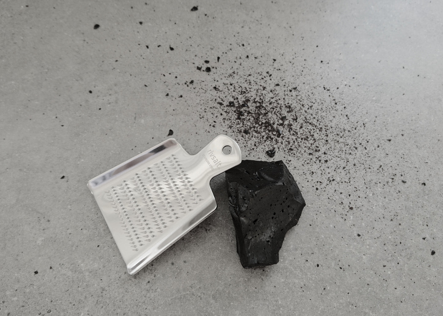 Rivsalt Raakalakritsi -tuotekuva; metalliraastin ja pala mustaa hiililiuskaa harmaalla alustalla, ympärillä hiilipulveria; raastin suorakulmainen, hopeanvärinen ja reiällinen; hiili matta ja kiinteä; ei tekstejä tai merkkejä; kuvakulma yläviistosta, ei pakkausta tai ihmisiä.