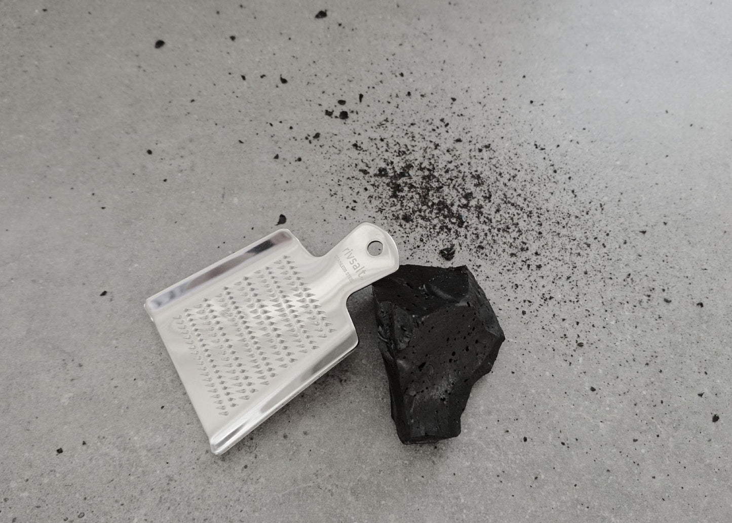 Rivsalt Raakalakritsi -tuotekuva; metalliraastin ja pala mustaa hiililiuskaa harmaalla alustalla, ympärillä hiilipulveria; raastin suorakulmainen, hopeanvärinen ja reiällinen; hiili matta ja kiinteä; ei tekstejä tai merkkejä; kuvakulma yläviistosta, ei pakkausta tai ihmisiä.