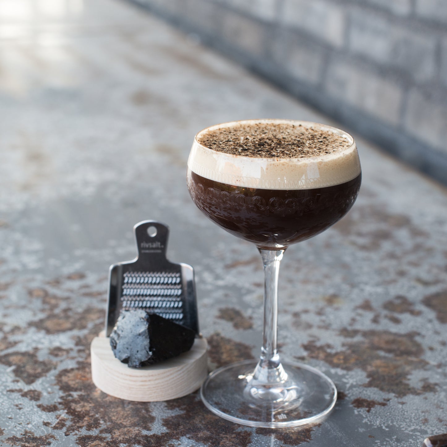 Rivsalt Raakalakritsi; coupe-lasillinen tummaa espresso martinia vaahdolla ja raastetulla mustalla tryffelillä päällystettynä, aseteltuna pienen suorakaiteen muotoisen metallisen raastimen ja epäsäännöllisen mustan tryffelinpalasen viereen tekstuurisella, kuluneella metallipinnalla; kuva lähikuva, ei näkyvää pakkausta tai luettavaa tekstiä, ei ihmisiä tai käsiä.