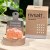 Rivsalt The Original Suolaraastin; metallinen raastin seisoo pyöreällä puisella alustalla, edessä pala vaaleanpunaista kivisuolaa, Rivsalt-brändätyn pahvilaatikon vieressä; laatikko ruskea tekstillä Rivsalt lihavoiduin mustin groteskikirjaimin; raastimessa soikea kahva ja rei'itetty litteä runko, molemmat mattametallia; taustalla vihreitä kasvinlehtiä; ei ihmisiä tai käsiä kuvassa.