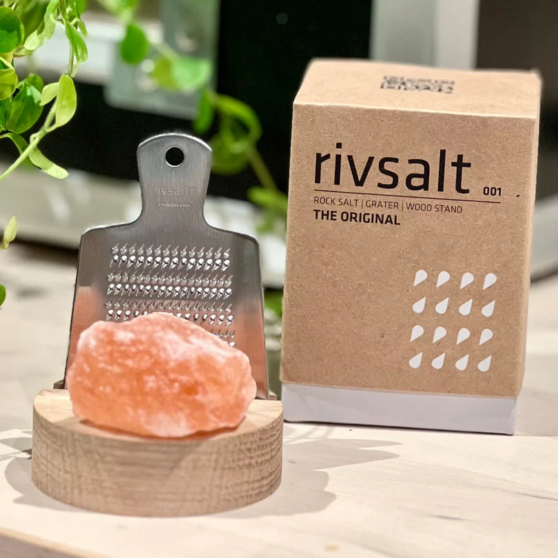 Rivsalt The Original Suolaraastin; metallinen raastin seisoo pyöreällä puisella alustalla, edessä pala vaaleanpunaista kivisuolaa, Rivsalt-brändätyn pahvilaatikon vieressä; laatikko ruskea tekstillä Rivsalt lihavoiduin mustin groteskikirjaimin; raastimessa soikea kahva ja rei'itetty litteä runko, molemmat mattametallia; taustalla vihreitä kasvinlehtiä; ei ihmisiä tai käsiä kuvassa.