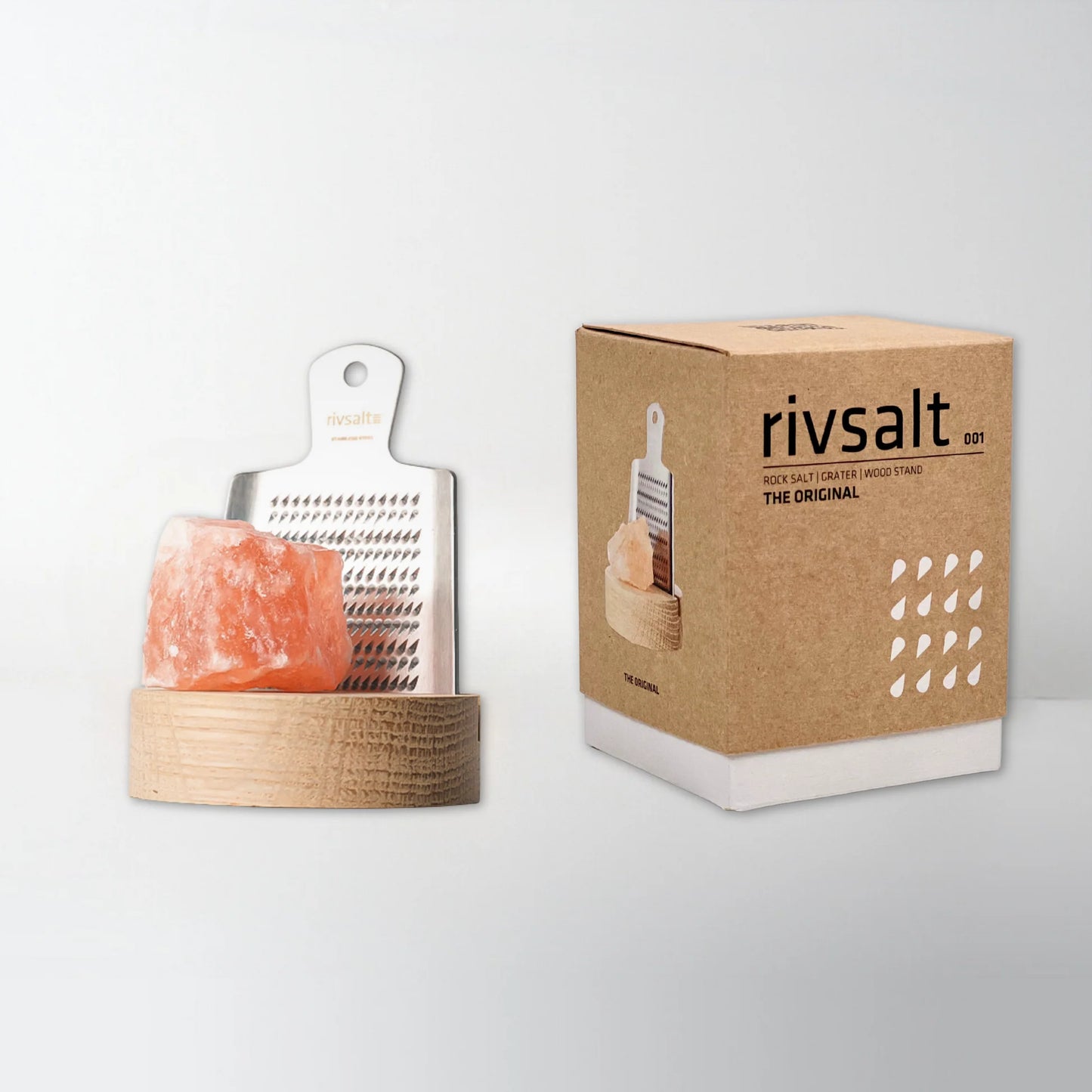 Rivsalt The Original Suolaraastin; Rivsalt-raastinsetti, jossa vaaleanpunaista kivisuolaa suorakaiteen muotoisella vaalealla puualustalla, aseteltuna pystyssä olevan mattapintaisen pahvipakkauksen viereen; laatikossa painettu tuotekuva ja teksti RIVSALT lihavoiduin groteskikirjaimin; tausta valkoinen; tuote ja pakkaus hyvin valaistuja, ei ihmisiä kuvassa.