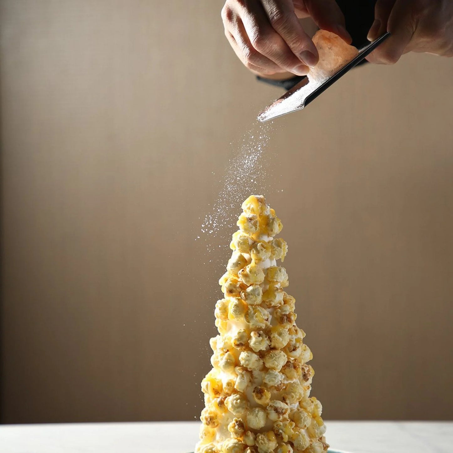 Rivsalt The Original Suolaraastin; käsi raastaa palaa Himalajan vaaleanpunaista suolaa kartionmuotoisen popcorn-tornin päälle neutraalia taustaa vasten; suolapala suorakaiteen muotoinen ja puoliläpikuultavan vaaleanpunainen; popcorn valkoista näkyvin pintarakentein, aseteltu kartiopinoon ilman pakkausta tai etikettejä; ei typografiaa näkyvissä; yksi valonlähde, lähikuva.