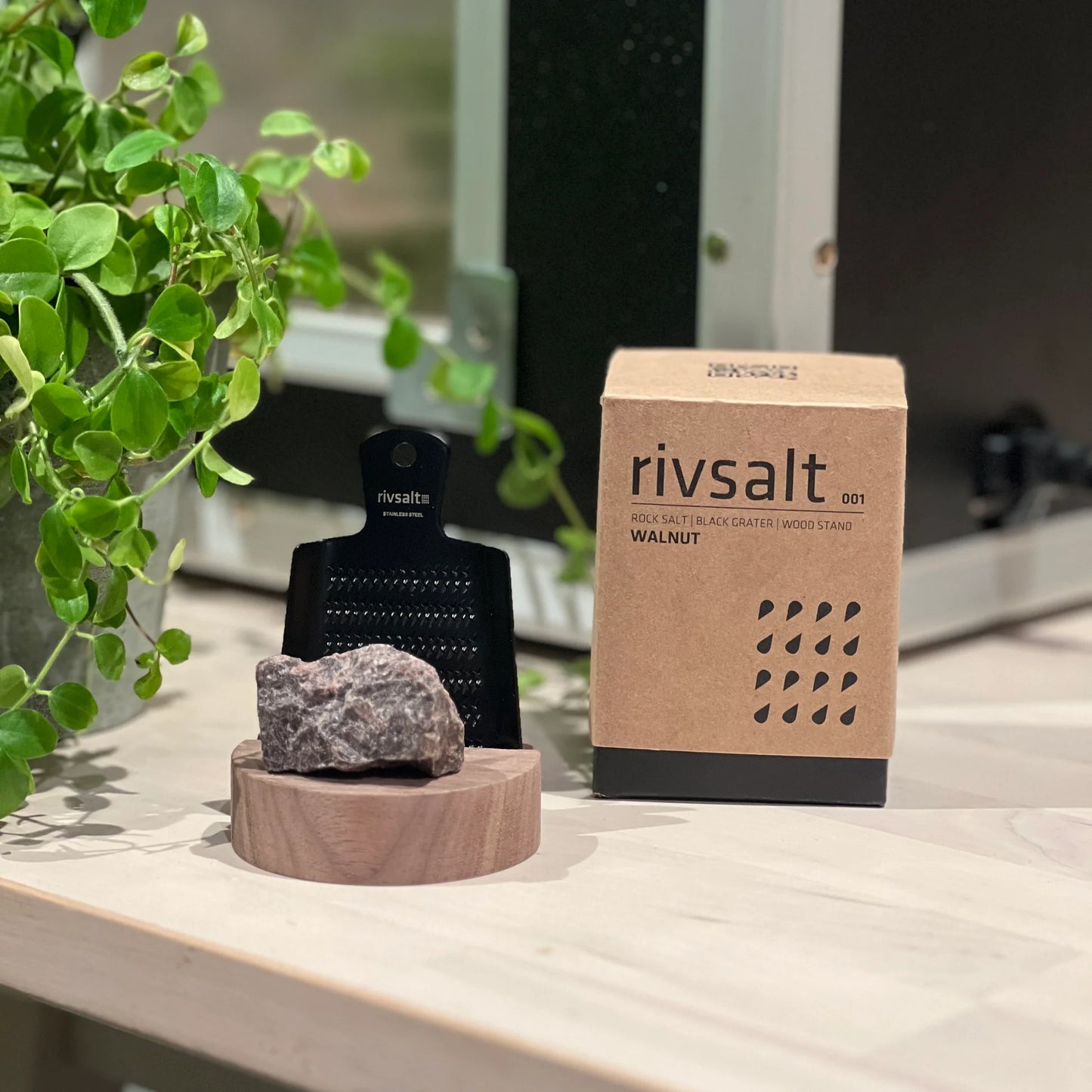 Rivsalt Walnut Suolaraastin; Rivsalt-kivisuolaraastinsetti mattamustalla metalliraastimella, yhdellä epäsäännöllisen muotoisella puoliläpikuultavalla kivisuolapalalla ja pyöreällä puisella alustalla, aseteltuna suorakaiteen muotoisen pahvipakkauksen viereen tekstillä rivsalt WALNUT lihavoiduin groteskikirjaimin; kaikki esineet tasaisella, vaalealla pinnalla vihreiden kasvinlehtien lähellä; ei ihmisiä tai käsiä kuvassa.