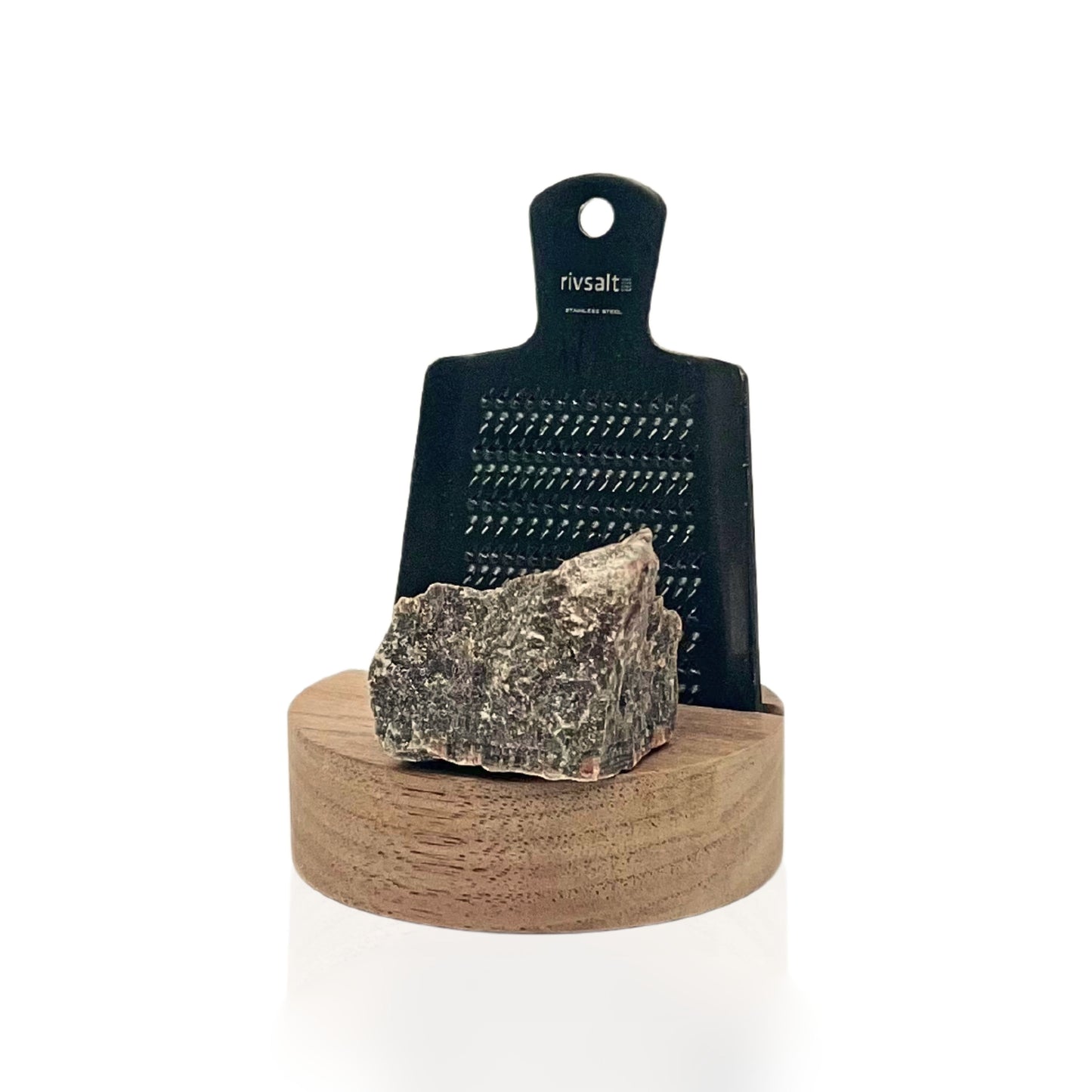Rivsalt Walnut Suolaraastin; metallinen raastin tekstillä rivsalt seisoo pystyssä pyöreällä puisella alustalla, edessä suuri pala kivisuolaa; ruostumattomasta teräksestä valmistettu raastin harjatulla pinnalla, suorakaiteen muotoinen pyöristetyin reunoin; teksti rivsalt painettu vaakasuunnassa minimalistisella groteskifontilla; pyöreä alusta vaaleaa puuta; ei pakkausta näkyvissä; tausta valkoinen, tuote keskitetty, ei ihmisiä kuvassa.