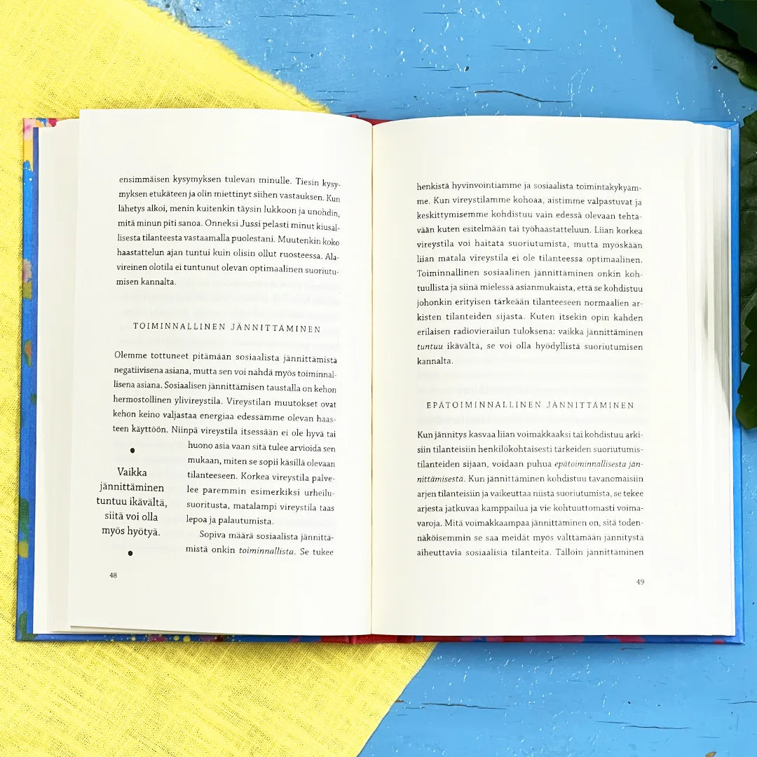 Rohkaisukirja ujoille; avoin kirja suomenkielisin tekstein lepää sinisellä ja keltaisella pinnalla; sivut mattapintaisia, mustaa antiikvatekstiä; ei kantta tai otsikkoa näkyvissä; alla oleva pinta tasainen, selkeät siniset ja keltaiset värikentät ilman tekstuuria; vihreä, leveä ja kiiltävä lehti tulee kuvaan oikealta reunalta; ei pakkausta tai ihmisiä näkyvissä.