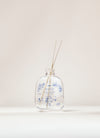 SEES x Moomin Room Diffuser Nordic Summer; kirkas lasinen diffuusoripullo sinisin kukkakuvioin, tekstillä SEES, sisältää tuoksunestettä ja kaksi ohutta tikkua, aseteltuna vaalealle taustalle; lasi sileää ja kiiltäväpintaista; pullon muoto lieriömäinen lyhyellä kaulalla; etiketissä SEES isoin groteskikirjaimin; ei ihmisiä kuvassa; kuva suoraviivainen ja hyvin valaistu.