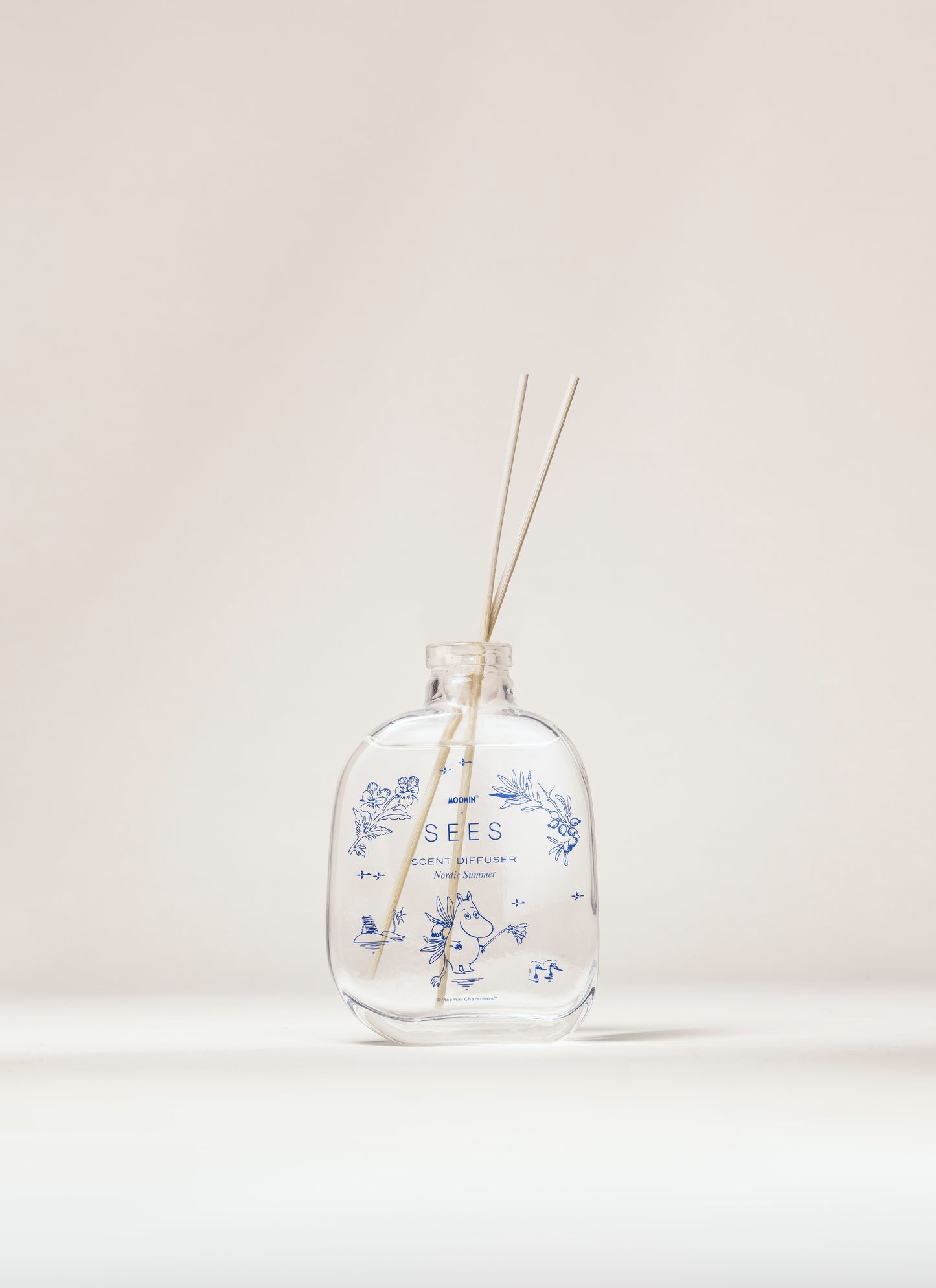 SEES x Moomin Room Diffuser Nordic Summer; kirkas lasinen diffuusoripullo sinisin kukkakuvioin, tekstillä SEES, sisältää tuoksunestettä ja kaksi ohutta tikkua, aseteltuna vaalealle taustalle; lasi sileää ja kiiltäväpintaista; pullon muoto lieriömäinen lyhyellä kaulalla; etiketissä SEES isoin groteskikirjaimin; ei ihmisiä kuvassa; kuva suoraviivainen ja hyvin valaistu.