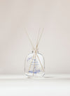 SEES x Moomin Room Diffuser Nordic Winter; kirkas lasinen tuoksudiffuusoripullo vaalein puutikuin ja hienovaraisin sinisin kasvikuvioin seisoo pystyssä neutraalia taustaa vasten; pullo lieriömäinen ja kiiltävä, valmistettu läpinäkyvästä lasista; ohuita, suoria tikkuja ulottuu aukosta; ei etikettiä tai tekstiä pinnalla; tausta tasaisesti valaistu ilman rekvisiittaa tai ihmisiä kuvassa.