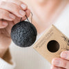 Saaren Taika Ecolution Aktiivihiili Konjac Puhdistussieni; henkilö pitelee mustaa pyöreää konjac-sientä narulla toisessa kädessä ja suorakulmaista pahvilaatikkoa toisessa; mattapintainen laatikko merkitty "Aktivihiili Konjac-puhdistussieni" lihavoidulla sans-serif-tekstillä; neutraali tausta; lähikuva keskittyy käsiin, tuotteeseen ja pakkauksen yksityiskohtiin.