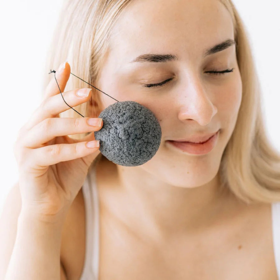 Saaren Taika Ecolution Aktiivihiili Konjac Puhdistussieni; vaaleahiuksinen henkilö silmät kiinni pitelee hellästi pyöreää mustaa konjac-sientä huokoisella tekstuurisella mattapinnalla lähellä poskeaan; ei näkyvää pakkausta tai etikettitekstiä; tausta valkoinen; lähikuva keskittyy käteen, sieneen ja osaan kasvoista.