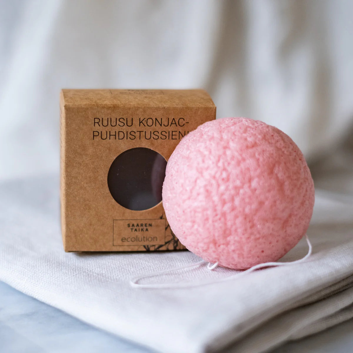Saaren Taika Ecolution Ruusu Konjac Puhdistussieni; vaaleanpunainen konjac-puhdistussieni valkoisella narulla taitellun kankaan päällä, vieressä ruskea suorakaiteen muotoinen rasia tekstillä ”Ruusukonjac-puhdistussieni” yksinkertaisella groteskifontilla; sieni pyöreä, hieman kupera muoto ja mattapinta; ei ihmisiä tai käsiä kuvassa; tausta pehmeästi sumennettu.