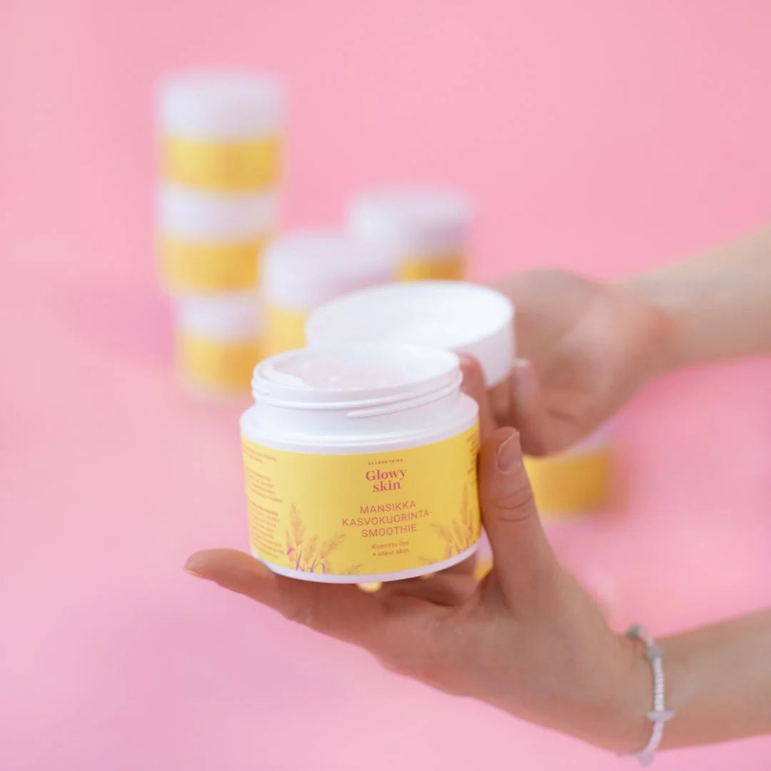Saaren Taika Glowy Skin Mansikka Kasvokuorinta Smoothie; henkilö pitelee avointa sylinterimäistä purkkia tekstillä "Glowy Skin" kasvokuorinnalle, matalla keltaisella etiketillä ja mustalla groteskitekstillä; purkki muovia valkoisella kannella, näyttää rakeista voidetta sisällä; useita suljettuja saman tyyppisiä purkkeja pinottuna ja epätarkkoina vaaleanpunaisella taustalla; lähikuva tuotteesta.
