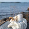 Saaren Taika Pyykkietikka Laventeli; pullo valkoisella etiketillä lepää paksukuteisen neulotun peiton päällä kuivattujen heinien kanssa, aseteltuna kivelle rauhallisen järven äärellä pilvisen taivaan alla; pullo sylinterimäinen ja suorareunainen, pakkaus läpikuultamaton tai puoliläpikuultava; etikettiteksti vähäistä ja pääosin mustaa groteskia; ei ihmisiä näkyvissä; ulkona luonnonvalossa, laaja kuva.
