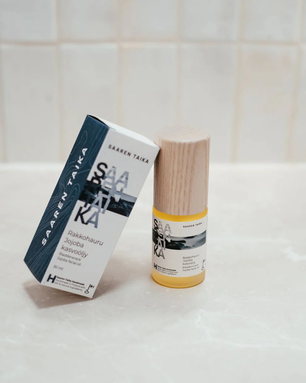 Saaren Taika Rakkohauru Jojoba kasvoöljy; pullo Saaren Taika -kasvoöljyä puisella korkilla lepää sinisen ja valkoisen vegaanisen ihonhoitopakkauksensa vieressä valkoisella laattapinnalla; pullo on sylinterinmuotoinen, lasia, mattapinnalla; puukorkki on sileä ja luonnonsävyinen; pakkauslaatikossa pelkistettyä sinistä tekstiä ”Saaren Taika” ja lisätietoja pienemmällä fontilla; ei ihmisiä tai käsiä näkyvissä.