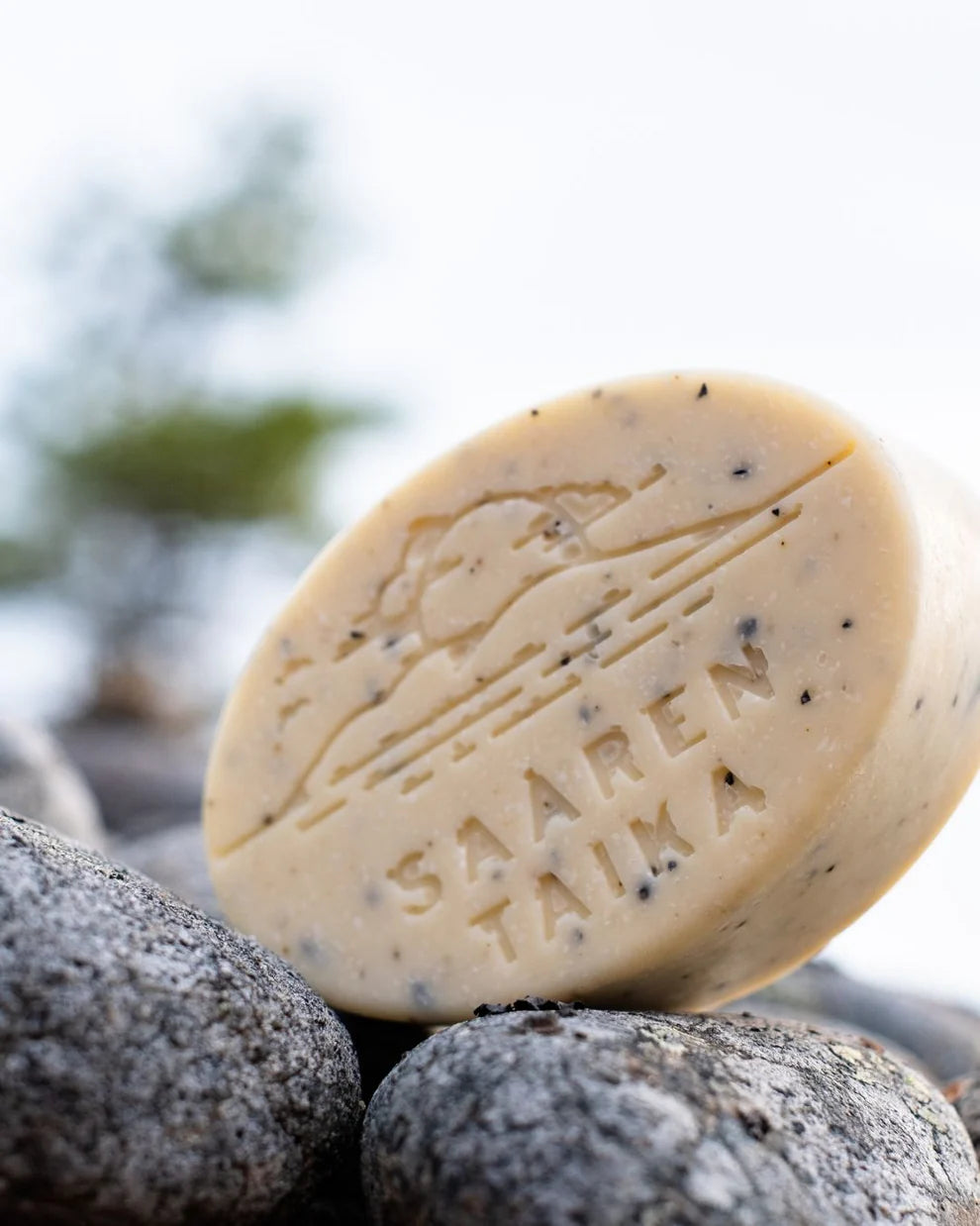 Saaren Taika Rakkohauru Suolasaippua; pyöreä beige saippuapala mattapinnalla, jonka yläosaan on kaiverrettu ”SAAREN TAIKA” ja luonnonaiheinen maisema; pakkausta ei näy; typografia on suuraakkosin; saippua lepää sileillä harmailla kivillä ulkona, taustalla epätarkkaa vihreää lehvästöä; ei ihmisiä tai käsiä näkyvissä.