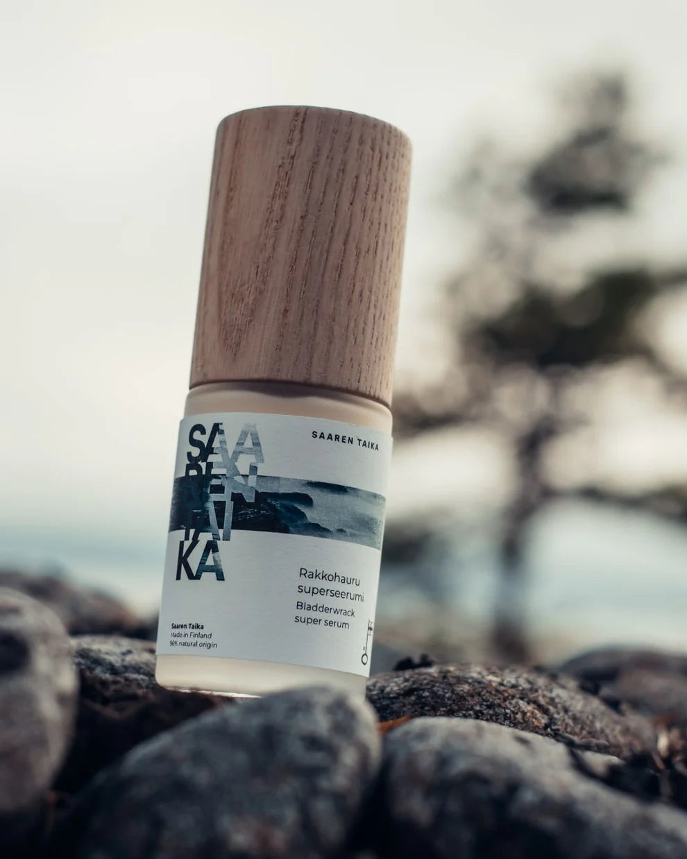 Saaren Taika Rakkohauru Superseerumi; pullo Saaren Taika Bladderwrack super serum -tuotetta, sylinterinmuotoinen läpinäkyvä runko ja vaaleanruskea puukorkki, seisoo pystyssä karkealla, epätasaisella kivipinnalla; pullon etiketti on valkoinen sinisin ja mustin groteskitekstein, kertoen tuotenimen; kivipinta on harmaa, taustalla epätarkka rannikkomaisema; ei ihmisiä tai käsiä näkyvissä.