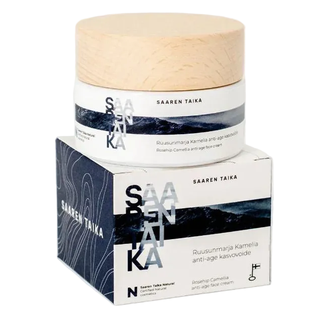 A jar of Saaren Taika Ruusunmarja Kamelia anti-age face cream sits atop its matching box, featuring minimalist white, blue, and wood-toned packaging with bold text and nourishing rosehip. The jar is cylindrical with a matte white finish and a light wood-colored flat lid. The label has bold black sans-serif text. The box repeats the same colors and fonts as the jar. Background is a seamless white surface. No people or hands are present in the image. This is a centered studio product shot at eye level.