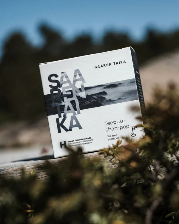 Saaren Taika Teepuu Shampoo, pakkaus ulkona puupinnalla, taustalla rehevää vihreyttä ja hiekkaista maisemaa pehmeästi epätarkkana; pakkaus suorakaiteen muotoinen mattapintaista pahvia, teksti yksinkertaista mustaa groteskia; ei ihmisiä eikä käsiä näkyvissä, tuote kuvattuna läheltä paikallaan olevana still-kuvana.