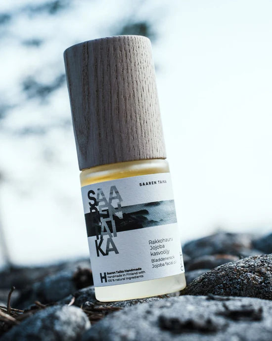 Saaren Taika Rakkohauru Jojoba kasvoöljy; lähikuva Saaren Taika -ihonhoitopullosta sylinterinmuotoisena, puisella korkilla, mattavalkoisella pinnalla ja mustalla antiikvatekstillä etiketissä; etiketissä yksinkertainen luontoaiheinen kuvitus, kuten lehtiä tai oksia; pullo on sijoitettu harmaalle tekstuuripintaiselle kivelle ulkona; ei ihmisiä tai käsiä näkyvissä; luettavassa tekstissä lyhyt lista hellävaraisia ainesosia.