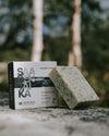 Saaren Taika Rakkohauru Kasvosaippua; täpläinen luonnonsaippuapala suorakulmaisella muodolla, mattapinnalla ja luonnonvalkoisella värillä seisoo yhteensopivan suorakulmaisen laatikon vieressä, jossa etikettiteksti ”Saaren Taika” yksinkertaisella groteskifontilla; molemmat esineet lepäävät karkealla kivipinnalla ulkona; taustalla epätarkkoja vihreitä puita; ei ihmisiä tai käsiä näkyvissä.