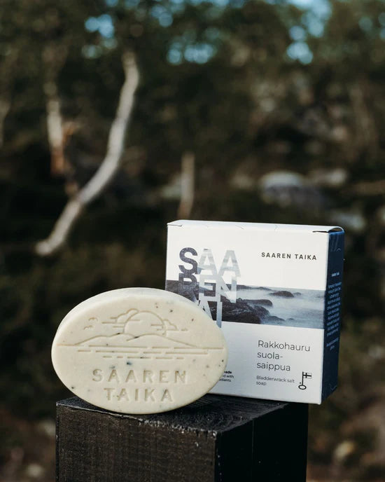 Saaren Taika Rakkohauru Suolasaippua; soikea saippuapala, merkitty Saaren Taika, sijaitsee laatikkonsa edessä, jossa luontoaiheinen maisemakuva; saippuassa sileä, mattapintainen viimeistely ja vaalea väri; laatikko seisoo pystyssä saippuan takana lohkotyylisin kirjaimin; kuva otettu ulkona, taustalla epätarkkoja vihreitä ja ruskeita puita; ei ihmisiä tai käsiä näkyvissä.
