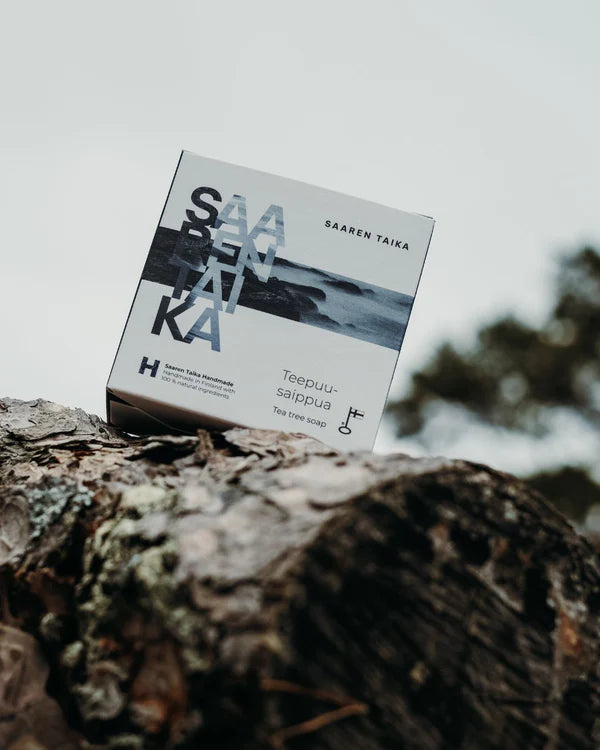 Saaren Taika Teepuusaippua; suorakulmainen mattapintainen pakkaus karkeasti sahatulla kannolla ulkona, teksti "Saaren Taika Tea Tree Soap" vihreällä ja harmaalla yksinkertaisella groteskikirjasimella pääosin valkoisella pohjalla; taustalla sumentuneita puita ja pilvinen taivas; ei ihmisiä tai käsiä kuvassa.