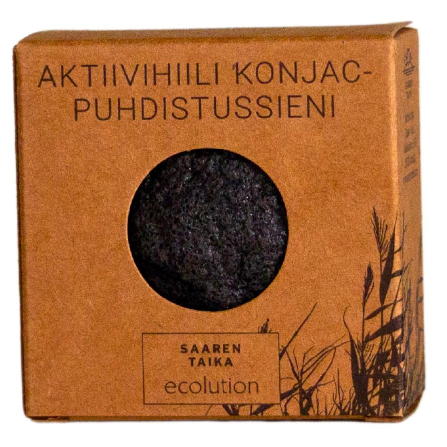 A brown box labeled Aktiivihiili Konjac-Puhdistussieni with a round black sponge visible through a circular window. Cardboard packaging has matte finish, rounded corners, and printed plant drawings. Text includes Saaren Taika Ecolution in mixed case sans-serif font. Box is photographed front-on against a neutral background. No people or hands are visible in the shot.