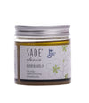 A jar of Sade Classic Kuorintaöljy exfoliating oil with a reflective silver lid, cylindrical shape, and transparent body showing pale yellow oil. The product label is in Finnish with dark green floral graphics on a white background, sans-serif fonts, and lists olive oil, peppermint oil, crystal salt. No people are present; neutral background with soft lighting.