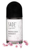 Sade Mineraalisuoladeodorantti Roll-On Ruusu; roll-on-deodorantti tekstillä "SADE crystal mineraalisuoladeodorantti Ruusu" mustalla korkilla, hienovarainen kukkakuvitus ja hehkuvia vaaleanpunaisia ruusunlehtiä ripoteltuna pullon juurelle; pullo lieriön muotoinen kiiltäväpinnalla; etiketti selkeällä modernilla typografialla; hyvin valaistu, ei ihmisiä tai käsiä; valkoinen tausta studiokuvassa.