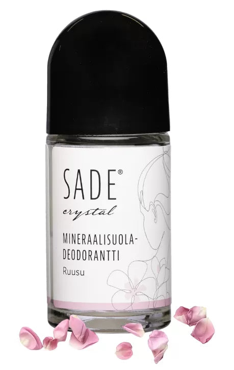 Sade Mineraalisuoladeodorantti Roll-On Ruusu; roll-on-deodorantti tekstillä "SADE crystal mineraalisuoladeodorantti Ruusu" mustalla korkilla, hienovarainen kukkakuvitus ja hehkuvia vaaleanpunaisia ruusunlehtiä ripoteltuna pullon juurelle; pullo lieriön muotoinen kiiltäväpinnalla; etiketti selkeällä modernilla typografialla; hyvin valaistu, ei ihmisiä tai käsiä; valkoinen tausta studiokuvassa.
