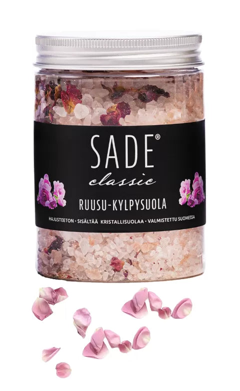 Sade Ruusu Kylpysuola; lieriön muotoinen kirkas lasipurkki valkoisella etiketillä tekstillä "SADE classic" mustalla sans-serif-fontilla, sisältää karkeaa vaaleanpunaista kylpysuolaa sekoitettuna näkyvien kuivattujen ruusunlehtien kera; vaaleanpunaisia ruusunlehtiä ja epäsäännöllisiä suolakiteitä ripoteltuna tasaiselle valkoiselle pinnalle purkin edessä; tausta valkoinen, ei ihmisiä tai käsiä.