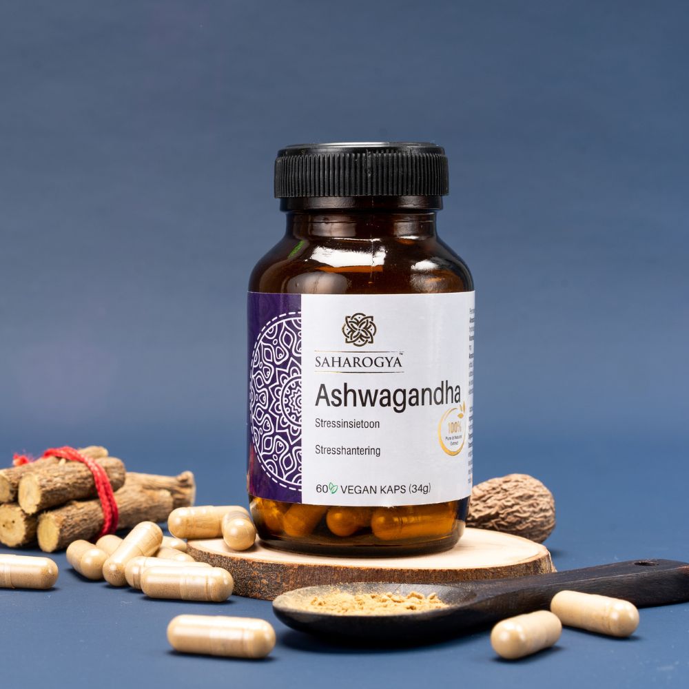 A bottle of Saharogya Ashwagandha capsules sits on a round wooden tray, surrounded by ashwagandha powder, capsules, and wooden sticks, set against a blue background. The bottle is plastic with a white screw cap and matte finish. Its green label features white and yellow text in sans-serif font reading “Saharogya Ashwagandha.” There are no people or hands visible. This is a studio shot.