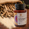 Pullo ashwagandhatabletteja seisoo kuivattujen ashwagandhajuurien ja jauhetun ashwagandhan edessä, taustalla osa kudottua säkkiä. Pullo on muovia, valkoinen ja lieriömäinen, kierrekorkillinen; etiketissä lukee Ashwagandha suurikokoisella pääteviivattomalla fontilla. Kohtaus on kuvattu edestä pöytäasetelmana. Ihmisiä tai käsiä ei näy.