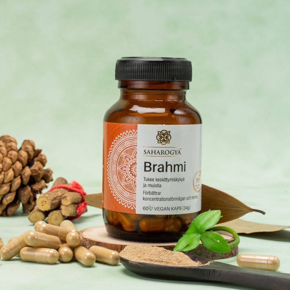 A bottle of Saharogya Brahmi capsules is positioned upright, surrounded by loose capsules, a mound of beige-brown powder, green leaves, and a pine cone. The bottle has a white plastic body with a green screw cap and matte finish. The label features clear sans-serif text stating its support for concentration, memory, and that it is vegan-friendly. Background: solid green. No people are present.