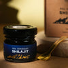 Saharogya Himalajan Shilajit pihka; pieni musta purkki tekstillä ”Aito Himalayan Shilajit” kultaisen lusikan ja vastaavan brändi-ilmeen ruskean suorakaiteen muotoisen rasian vieressä; molempien musta etiketti valkoisella groteskitekstillä; kaikki esineet vaalealla tasaisella alustalla pehmeässä valaistuksessa; ei ihmisiä tai käsiä kuvassa.