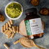 A bottle of Saharogya Karela capsules with a white plastic body and green cap features green and black printed label text. The bottle is placed on a matte, textured surface alongside a small bowl containing fine green powder, loose tan capsules, several sliced pieces of fresh karela (bitter melon), and a metal spoon holding more karela powder. No people or hands are present. A bottle of Saharogya Karela capsules is surrounded by green powder in a bowl, tan capsules, sliced karela pieces, and karela powder