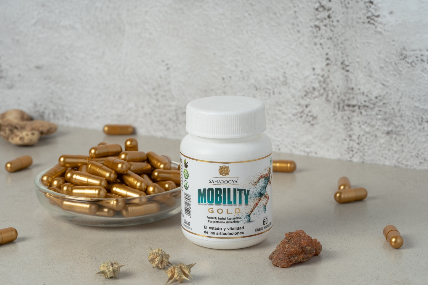 A bottle labeled Mobility Gold sits next to a bowl and scattered golden capsules, enriched with natural ingredients, alongside stones and shells on a beige surface against a textured background. Brown cylindrical plastic bottle with a matte finish; white cap; front label in simple black sans-serif text, “Mobility Gold” is prominent. Capsules are glossy oblong gold color. Ceramic bowl is white. No people or hands visible. Stones and seashells arranged around products on flat matte surface; full product