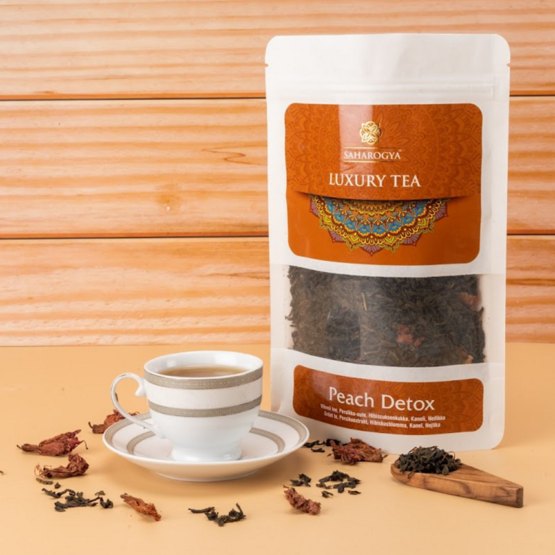 Teekuppi valkoisella lautasliinalla on aseteltu suorakaiteen muotoisen paperipakkauksen viereen, jossa lukee Saharogya Luxury Tea Peach Detox painetulla lihavoidulla serif- ja käsialatyylisellä fontilla; irtoteelehtiä ja kuivattuja terälehtiä on levitetty vaalean puupinnan päälle; tausta myös vaaleaa puuta; ei ihmisiä tai käsiä näkyvissä; keskikokoinen tuotekuva suoraan edestä.