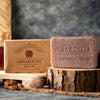 A bar of Saharogya Red Sandal wellness soap sits on a wooden slab with its box behind it. The soap is rectangular with rounded edges and has a reddish-brown color and a matte finish. The box features the brand name in clear, modern sans-serif font. Natural ingredients, including nourishing resin and a healing nut, are displayed nearby. The background features a rustic, spa-like setting.