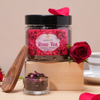 Saharogya Rose Tea Musta Tee; kirkas lasipurkki tekstillä ”Rose Tea” sisältää ruusunterälehtiä; tuore punainen ruusu, pieni läpinäkyvä lasikulho teetä, vaalea puinen kauha ja pelkistetty valkoinen teekuppi aseteltuna suorakaiteen muotoiselle mattapintaiselle vaalealle puupalikalle; tausta yksivärinen beige; esineet käyttämättöminä ja pystyssä; ei ihmisiä tai käsiä näkyvissä.