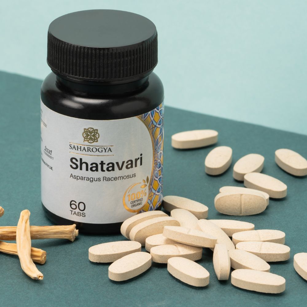A bottle labeled Shatavari sits on a flat surface with scattered oval tablets and a few dried roots, set against a teal and beige background. The bottle is cylindrical with a matte white finish and has a label reading “Shatavari” in clear sans-serif font. Tablets are light brown, oval-shaped; dried roots are fibrous and tan. No people or hands are present.