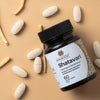 A black, matte-finished bottle sits angled on a beige surface. The label with “Shatavari” in clear, modern font is fully visible. White oblong tablets and pieces of dried Shatavari root are scattered nearby. No people or hands are present in the shot. Product and contents are arranged for clear visibility.
