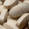 Close-up of several beige, oval-shaped tablets or supplements, stacked and overlapping each other. The textured surface of the vitamins is slightly rough. No visible packaging or labels. Tablets have a matte, uneven finish. No typography present in the image. Plain, neutral background with no discernible setting details. No people or hands visible in the frame.