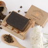 A rectangular dark soap bar is on a wooden, slatted soap dish next to a matte-finish brown rectangular box with “Saharogya Wellness Soap” in clear, sans-serif font. Small loose seeds are scattered near an upside-down ceramic cup with a light glaze. A single white flower rests nearby. All objects are precisely arranged on a solid matte white surface. No people or hands present.