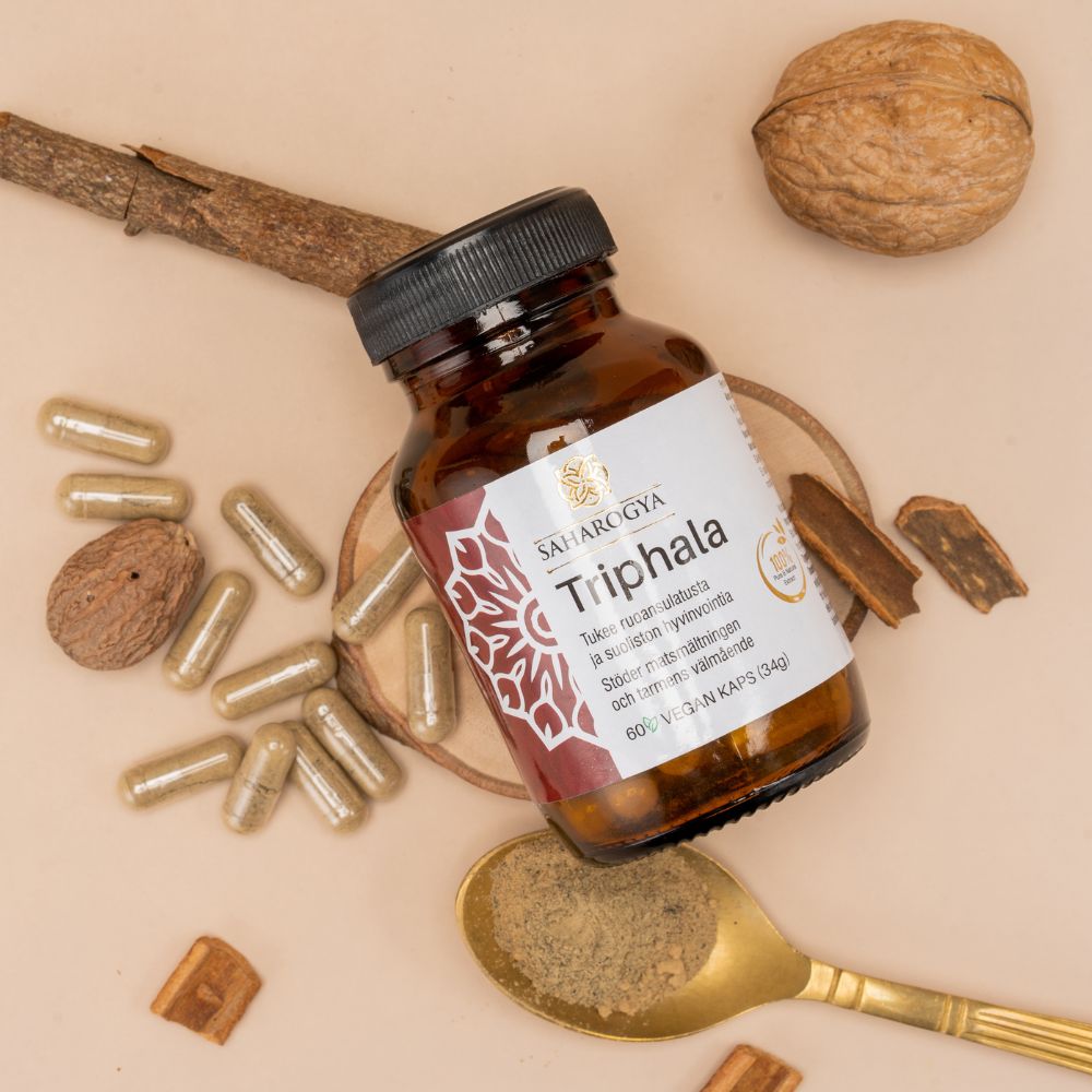 A bottle labeled Triphala lies on a surface with capsules, a gold spoon filled with powder, a walnut, cinnamon sticks, nutmeg, and Ayurvedic ingredients nearby. The bottle is cylindrical and made of opaque material with a matte label that shows bold uppercase text reading “Triphala.” The shot is flat-lay style on a neutral-colored background. No people or hands are visible.