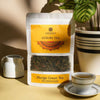 Saharogya Mango Green Tea; mattapintainen suorakulmainen pussi sileällä beigellä pöytäpinnalla; lähellä valkoinen posliinikuppi höyryten, kiiltävä valkoinen maitokannu ja vihreä ruukkukasvi taustalla; kaikki esineet kirkkaasti valaistu auringonvalossa; kuvassa ei ole ihmisiä tai käsiä.