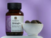 Saharogya Shilajit+ tabletti; pullo tekstillä ”Shilajit+” pienen valkoisen kulhon vieressä, jossa pyöreitä, tummia shilajit-tabletteja, vaaleanvioletilla taustalla; pullo lieriömäinen ja yksivärisestä materiaalista mattapinnalla, niukkaa groteskityyppografiaa; molemmat esineet tasaisella alustalla, ei ihmisiä tai käsiä; kuvaus yksinkertainen ja tasaisesti valaistu.