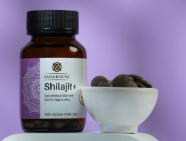 Saharogya Shilajit+ tabletti; pullo tekstillä ”Shilajit+” pienen valkoisen kulhon vieressä, jossa pyöreitä, tummia shilajit-tabletteja, vaaleanvioletilla taustalla; pullo lieriömäinen ja yksivärisestä materiaalista mattapinnalla, niukkaa groteskityyppografiaa; molemmat esineet tasaisella alustalla, ei ihmisiä tai käsiä; kuvaus yksinkertainen ja tasaisesti valaistu.